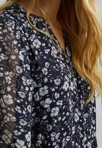 Femme aux longs cheveux blonds portant un chemisier bleu foncé avec de petits motifs floraux blancs et orange, boutonné à l'avant.