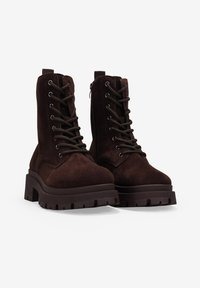 Botas de tobillo de ante marrón con una suela de goma gruesa, con cierre de cordones en la parte delantera, cremallera lateral y detalles texturizados en la parte superior.