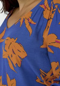 Blaue Stoffbluse mit großen orangefarbenen Blumenmustern und schwarzen Konturen. V-Ausschnitt-Design und lockere Passform mit gerafften Details an den Schultern.