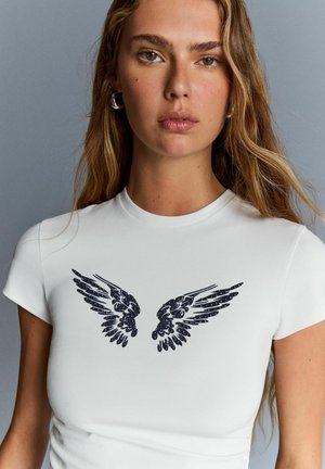 Woman with long hair wearing a white fitted t-shirt with black wing designs across the chest, standing against a gray background.