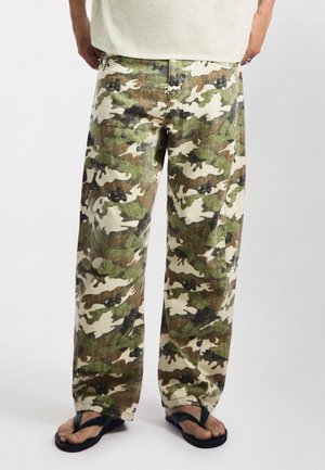 Personne portant un pantalon camouflage ample et des tongs noires debout sur un fond blanc.