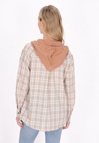 Geruite shirt in beige en bruin, met een bruine capuchon. De stof is gestructureerd met een zachte afwerking, en de onderkant is iets gebogen.