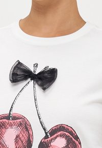 Camiseta blanca con un gráfico de cereza rosa y un lazo de malla negra como adorno en el escote. Tela suave con cuello redondo.