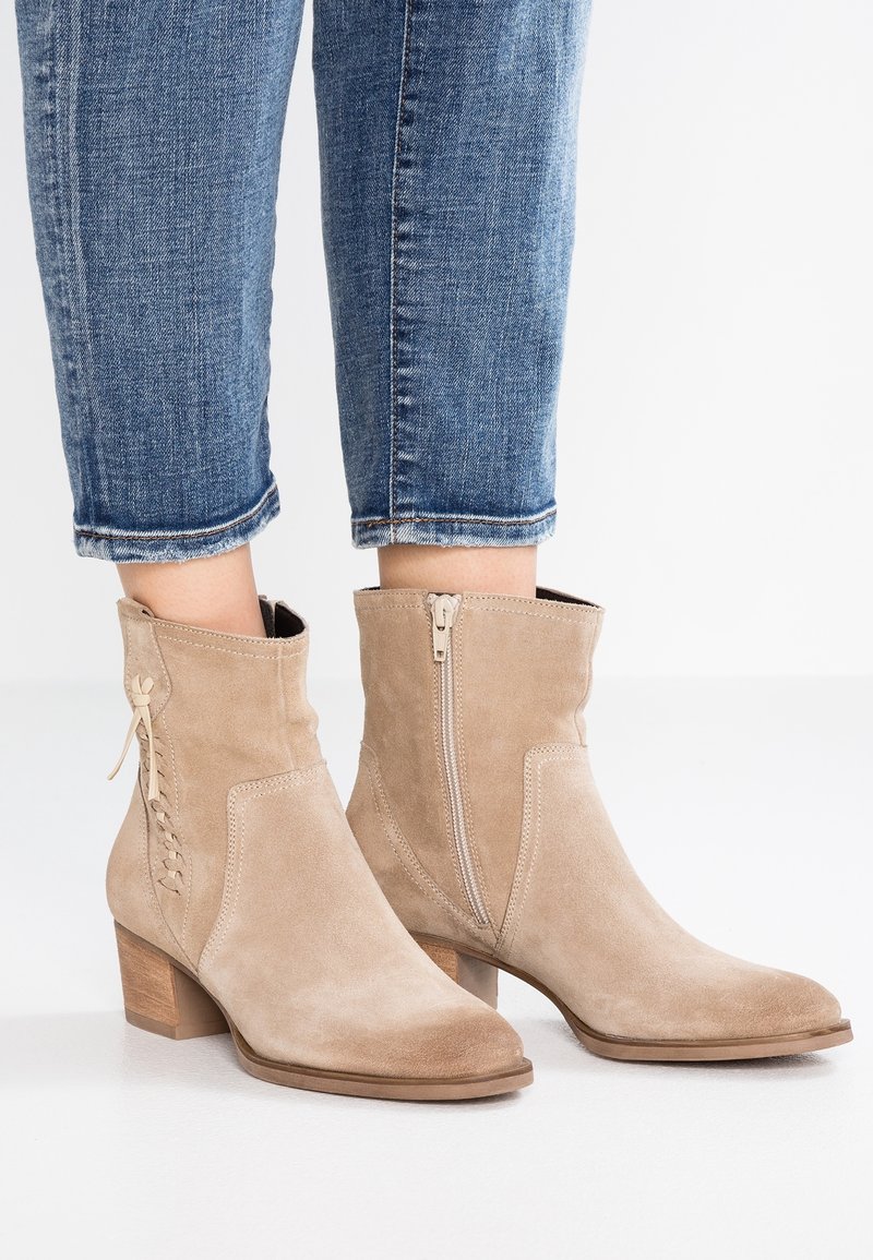 Beżowe zamszowe botki za kostkę z niskim, podniesionym obcasem, z bocznym zamkiem błyskawicznym i subtelnymi koronkowymi detalami po jednej stronie. Gładka tekstura i spiczaste noski.