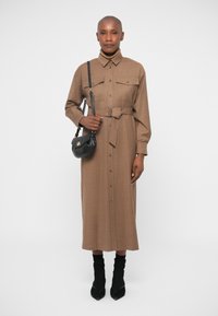 Καφέ φόρεμα πουλόβερ με μακριά μανίκια, κούμπωμα με κουμπιά και δύο μπροστινές τσέπες. Περιλαμβάνει και ζώνη στη μέση. Φορεμένο με μαύρες μυτερές γόβες και τσάντα.