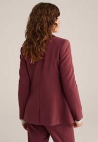 Žena s dlouhými kudrnatými hnědými vlasy, oblečená v přizpůsobeném tmavě fialovém saku a odpovídajících kalhotách, stojící bokem na béžovém pozadí.