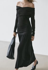 Woman wearing a long black off-shoulder dress with pleated skirt, carrying a black leather handbag, and wearing black heels.