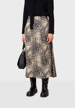 Femme portant une jupe midi imprimé léopard, un top noir à manches longues, des bottes noires, tenant un sac à main noir sur un fond uni.