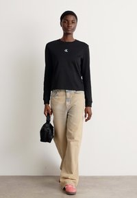 Schwarzes Langarmshirt mit kleinem blauen Logo, kombiniert mit beigen weitgeschnittenen Jeans und pinken Turnschuhen. Eine schwarze Handtasche vervollständigt das Outfit.