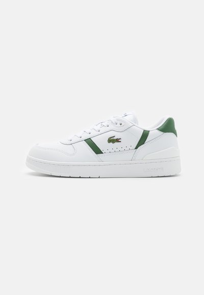 Бяла кожена маратонка със зелени акценти, логото на крокодила на Lacoste отстрани, и дебела подметка, показана от външната страна на едноцветен фон.