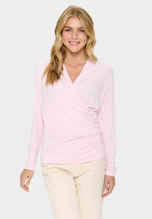 Haut rose à manches longues en tissu doux, avec un décolleté en V, des détails froncés sur le côté et des manches ajustées. Associé à un pantalon de couleur claire.