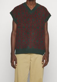 Tjock stickad tröja i mörkgrön med röd utsmyckad mönster. V-ringad design, korta ärmar och ribbad nederkant. Minimalistisk etikett synlig.