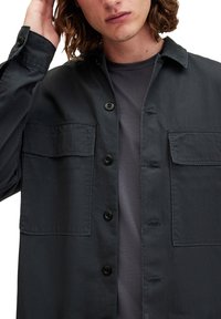Ciemnoszara koszula zapinana na guziki, wykonana z teksturowanej tkaniny, z dwoma przednimi kieszeniami, czarnymi guzikami i luźnym krojem na ciemnoszarym t-shircie.