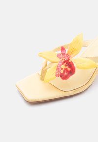 Μπεζ σανδάλι με τετράγωνη μύτη και μεγάλο κίτρινο και κόκκινο διακοσμητικό άνθος ορχιδέας στο λουράκι.