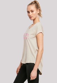 Blonde woman in beige short-sleeve shirt with pink text, wearing black leggings, standing sideways against plain light background.
