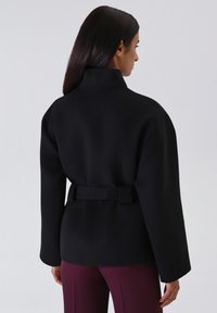 Cappotto nero con cintura, colletto alto, maniche ampie e texture liscia, abbinato a pantaloni plissettati color bordeaux. La vista posteriore mostra la forma del cappotto.