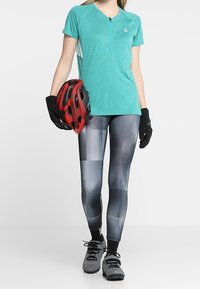 Tyrkysový krátký rukáv, zipový top v kombinaci s legínami s černobílým vzorem, rukavicemi a červenou cyklistickou helmou. Texturovaná látka.