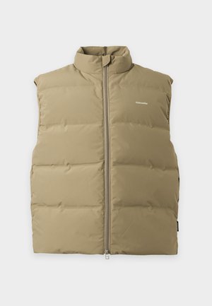 Personne portant un gilet beige matelassé sur un t-shirt noir à manches longues, un pantalon gris ample et des chaussures marron, debout devant un fond blanc uni.