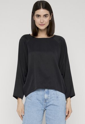 Femme aux cheveux raides et foncés, portant un haut noir à manches longues ample et un jean taille haute bleu clair, debout devant un fond uni.