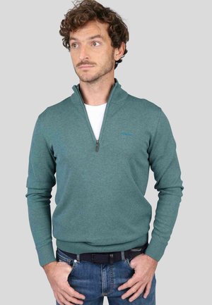 Pull en polaire teal avec un col montant, fermeture zippée mi-longue, poignets et ourlet côtelés. Présente un accent logo subtil. Porté par-dessus une chemise blanche.