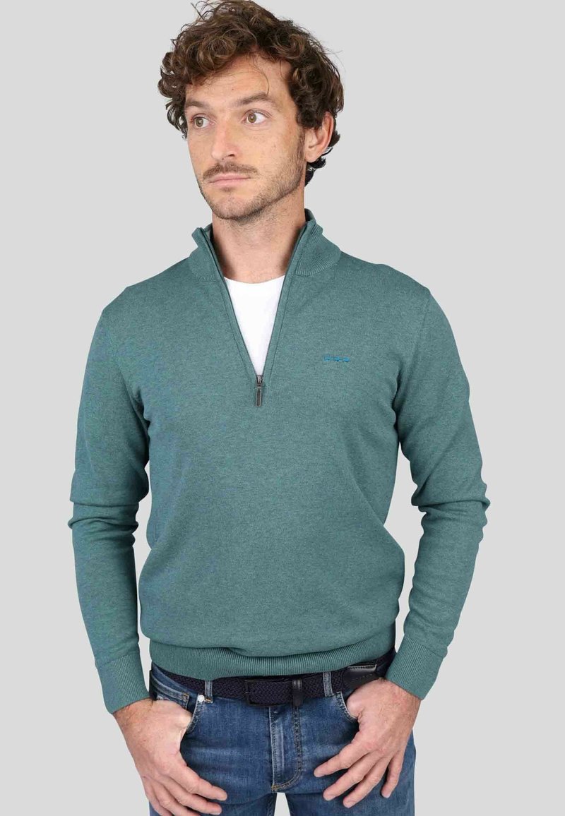 Pull en polaire teal avec un col montant, fermeture zippée mi-longue, poignets et ourlet côtelés. Présente un accent logo subtil. Porté par-dessus une chemise blanche.