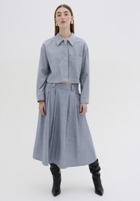 Pilkas chambray trumpas marškinėliai su apykakle ir kišene, derinami su raukšlėta midi sijonu. Modelis vilki juodus iki kelių batus. Minimalistinis dizainas.
