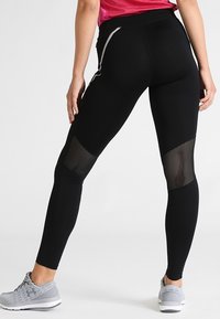 Černé sportovní legíny s přiléhavým střihem, texturované síťované panely na lýtkách a reflexní detaily. V kombinaci se šedými teniskami.
