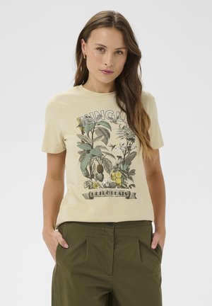Kobieta z długimi brązowymi włosami, ubrana w beżowy t-shirt "Jungle Retro Beats" z zielonymi ilustracjami roślin, w połączeniu z oliwkowozielonymi spodniami, ręce w kieszeniach.