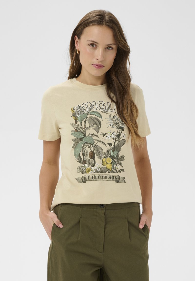 Femme aux longs cheveux bruns portant un t-shirt beige "Jungle Retro Beats" avec des illustrations de plantes vertes, assorti à un pantalon vert olive, les mains dans les poches.