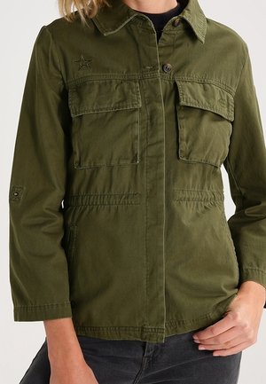 Kurtka zapinana na guziki w kolorze oliwkowej zieleni, z dwoma kieszeniami na piersi, wyszyciem w kształcie gwiazdy oraz elastycznym detalem w pasie, noszona na czarnym topie.