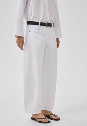 Egy személy fehér bőszárú nadrágot, fehér inget, fekete övet és fekete papucsot visel, sík háttér előtt áll.