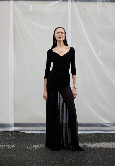 Rochie neagră cu mâneci lungi, design tip petrecut și fustă femeiască, stratificată și transparentă. Materialul este neted, cu o textură fluidă și o siluetă care se mulează pe corp.