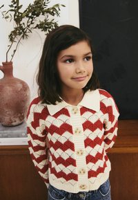 Cardigan à motifs rouge et crème avec un design à bord festonné, boutonnière sur le devant et un col, associé à un jean bleu. Manches courtes.