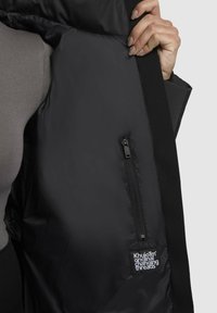 Crna puf jakna iznutra s glatkom teksturom, s patentnim zatvaračem i malom oznakom s logotipom. Materijal se čini izdržljivim i modernim.