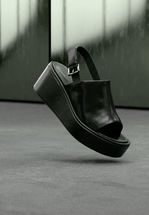 Sandale à plateforme en cuir noir avec une large bride, orteil ouvert et boucle de cheville réglable, montrée flottant au-dessus d'une surface grise.