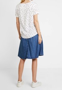 Camisa blanca de manga corta con lunares negros, combinada con una falda de mezclilla azul y zapatillas blancas, vista desde atrás.