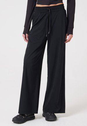 Personne portant un pantalon noir ample à cordon de serrage et des baskets noires, debout les mains détendues le long du corps devant un fond uni.