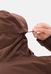 Veste imperméable marron avec une capuche ajustée. Un cordon de serrage est en cours d'ajustement, mettant en valeur la texture lisse du matériau et les détails des coutures.