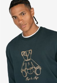 Donkergroen sweatshirt met een ronde hals. Voorzien van een gouden geborduurd teddybeerontwerp op de voorkant, met gedetailleerde gezicht- en lichaamssteken.