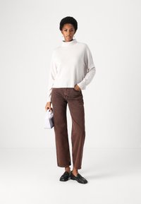 Jasnoróżowy sweter z golfem, brązowe szerokie dżinsy, czarne loafersy, trzymający małą fioletową torebkę. Prosty i czysty design.
