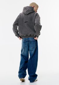 Hanorac gri cu glugă, cu o textură periată și un logo discret pe spate, asociat cu o pereche de blugi albaștri, de tip lejer, purtați într-un stil casual.