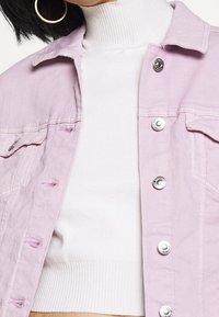 La chaqueta de mezclilla lila cuenta con botones a presión plateados, bolsillos frontales y un corte relajado, superpuesta sobre un suéter de cuello alto de canalé blanco.