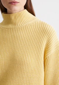 Żółty sweter dzianinowy z wysokim prążkowanym kołnierzem, o luźnym kroju i fakturowanym wzorze. Materiał wydaje się miękki i przytulny.