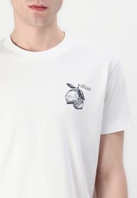 Balti medvilnės marškinėliai trumpomis rankovėmis su juodu piešiniu, vaizduojančiu apelsiną ir citriną su žalios lapeliais bei etikete "Super Jeans."