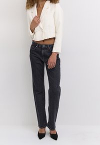 Weiße, kurz geschnittene Jacke aus strukturiertem Stoff, kombiniert mit dunklen, geraden Jeans. Das Model trägt schwarze spitz zulaufende High Heels.