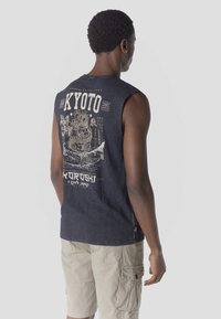 Hombre con camiseta sin mangas azul marino con la palabra "KYOTO" y un gráfico de dragón, combinado con pantalones cortos tipo cargo beige, de pie contra un fondo liso.