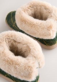 Grønne strikkede hjemmesko med en blød, beige faux fur-krave, rund form og tekstureret overflade med gule syninger.