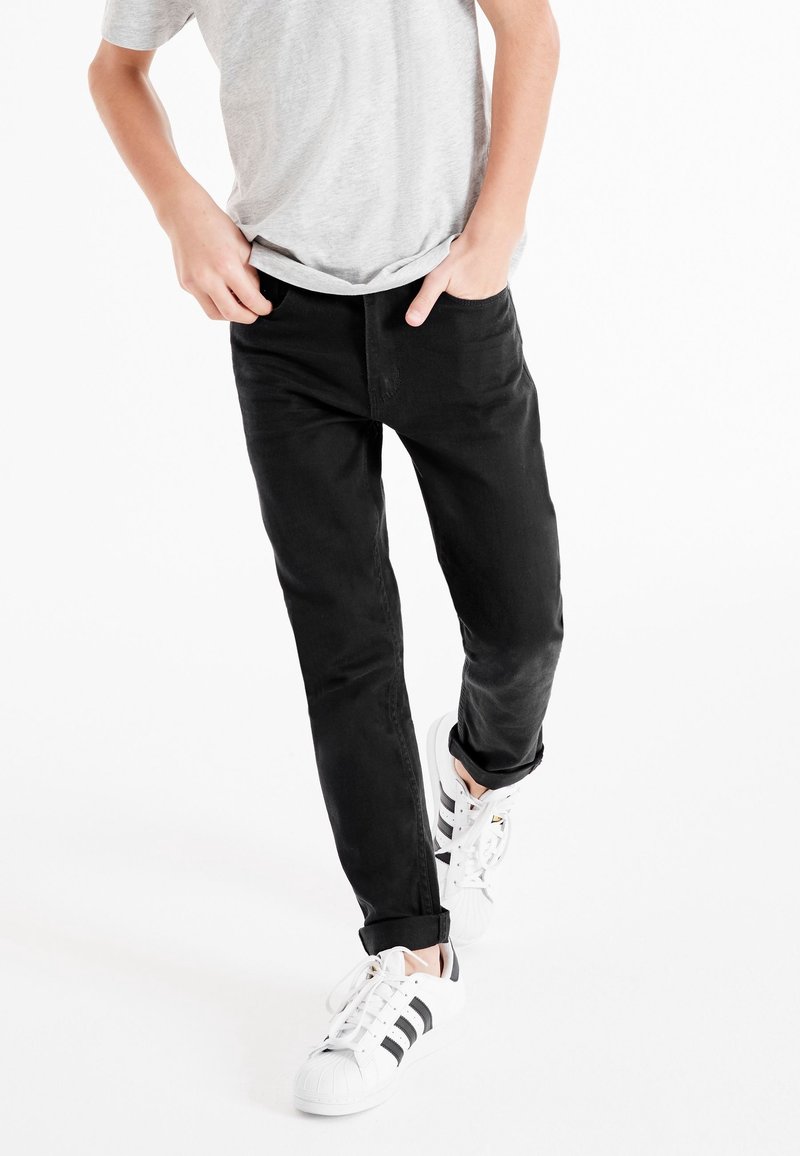 Personne portant un jean noir retroussé aux chevilles, un t-shirt gris clair et des baskets blanches avec des rayures noires, debout les mains dans les poches.