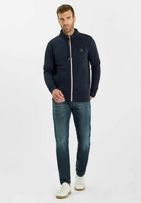 Marineblauw zip-up sweatshirt met opstaande kraag, lichtgewicht stof, witte ritssluiting en trekkoorden, gecombineerd met blauwe spijkerbroek en witte sneakers.