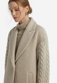 Beige ullkappa med skräddarsydd design, med kabelstickade ärmar och figurskuren krage. Strukturen av tyget och detaljer med sidofickor synliga.